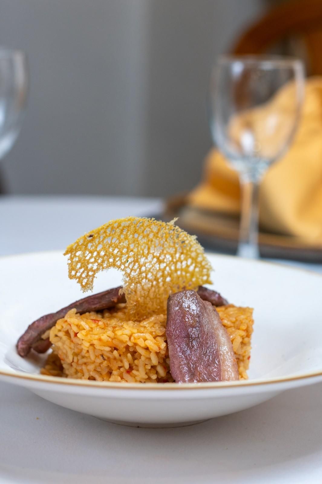 Plato gourmet de arroz con carne sellada y una guarnición crujiente sobre una mesa blanca, con copas de vino al fondo.