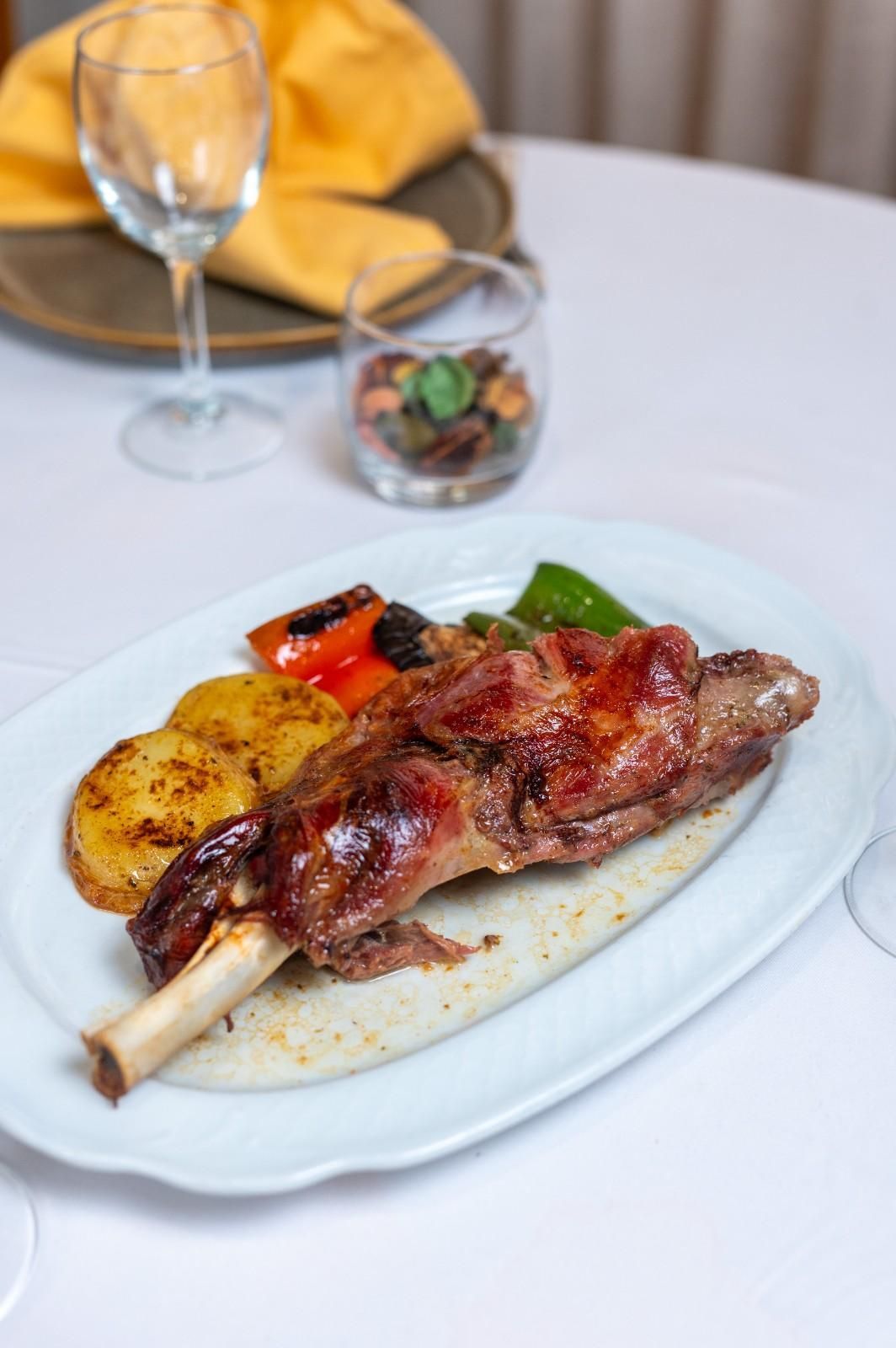 Carne a la parrilla con verduras asadas en un plato blanco sobre una mesa de comedor, con una copa de vino y pan al fondo.