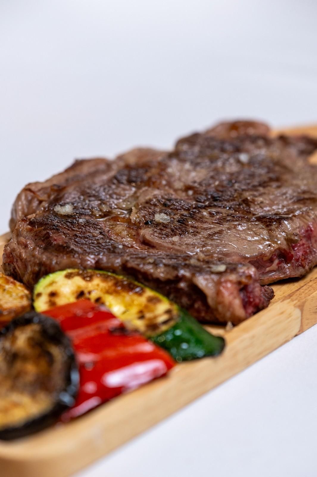 Filete a la parrilla sobre una tabla de madera con verduras asadas sobre un fondo blanco.