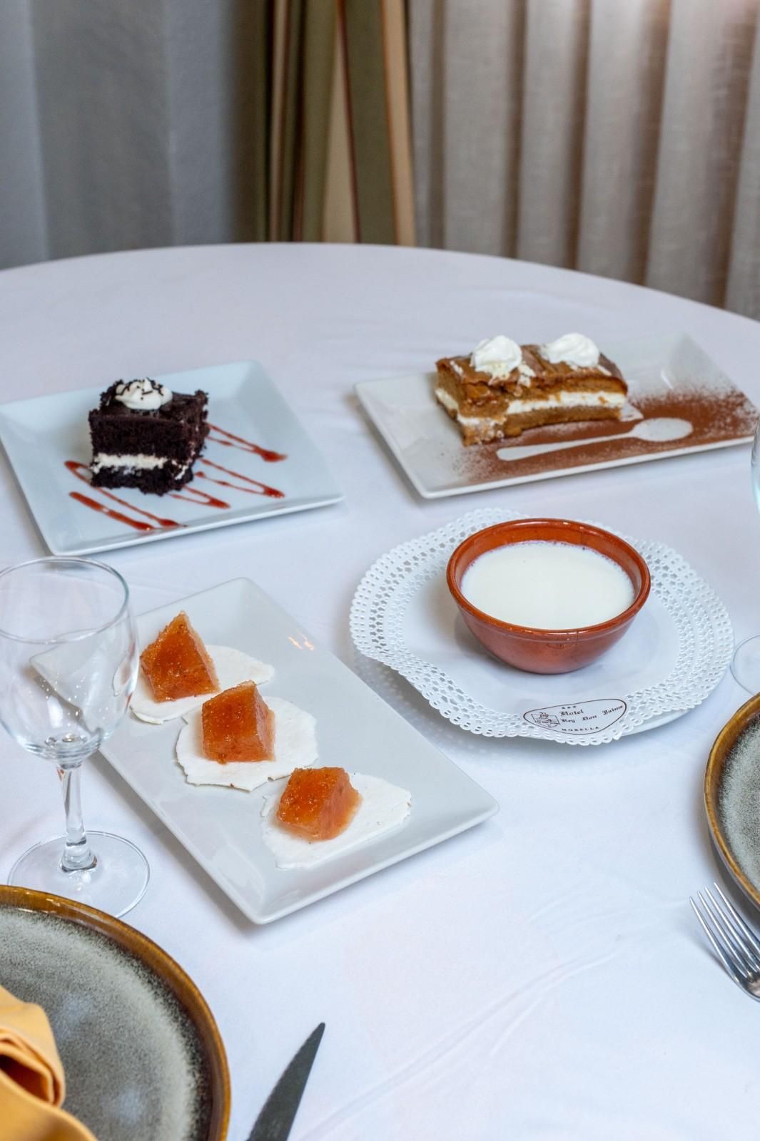 Mesa de postres con porciones de pastel, tartaletas y copas sobre platos blancos y un mantel blanco.