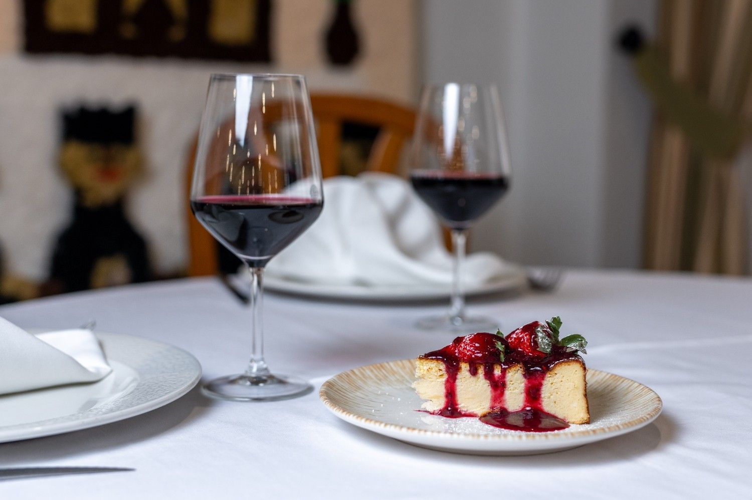 Plato de postre con salsa de frutos rojos y fresas junto a dos copas de vino tinto en una mesa puesta.