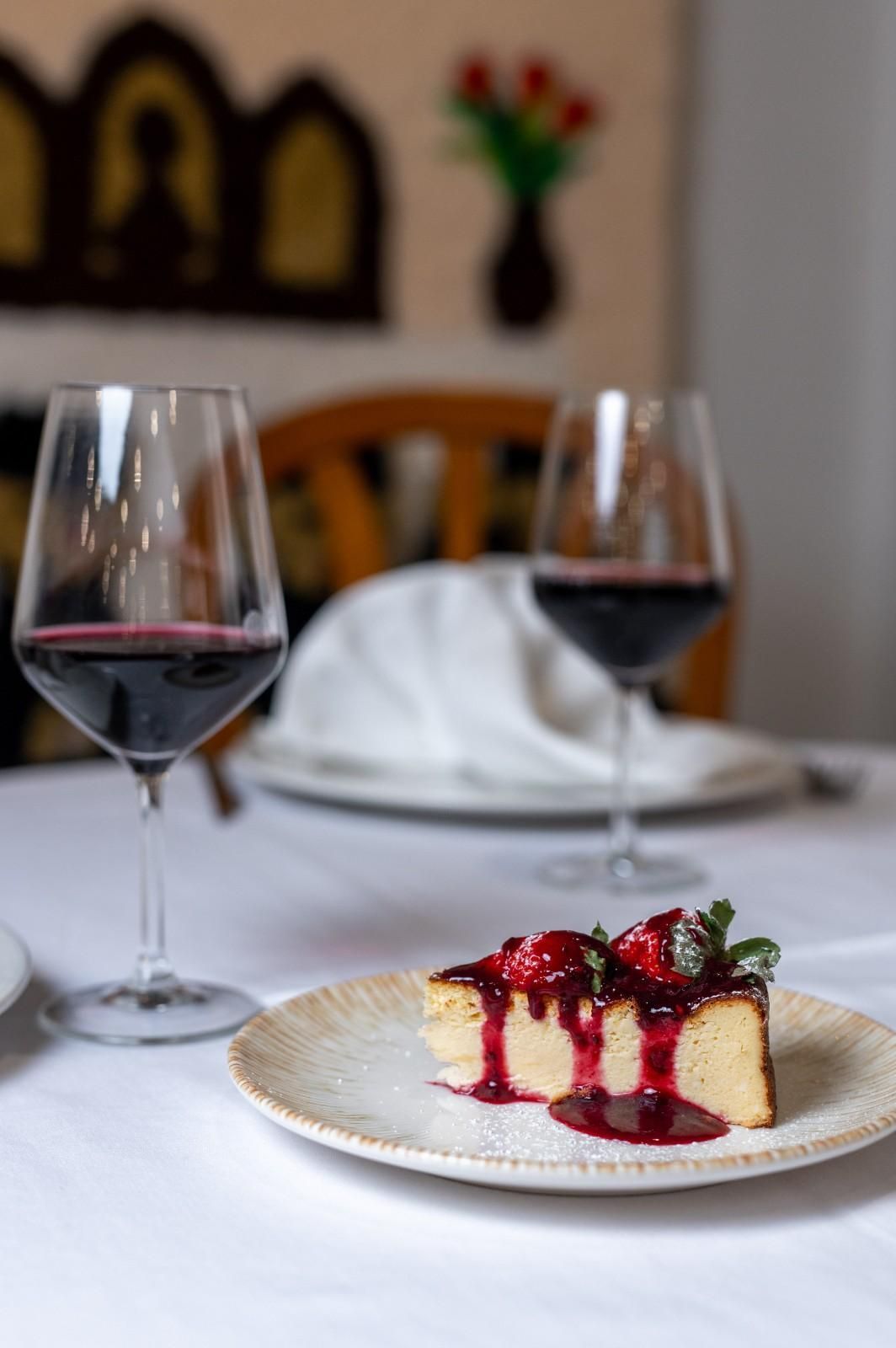 Una porción de tarta de queso con frutos rojos en un plato con dos copas de vino tinto en una mesa de comedor.