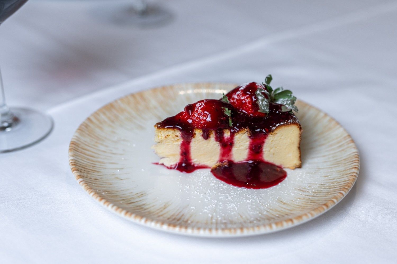 Porción de tarta de queso cubierta con salsa de frutos rojos en un plato decorativo, sobre un mantel blanco.