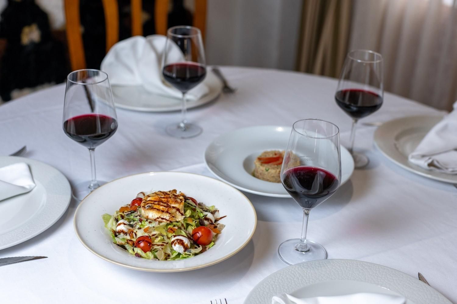 Elegante mesa de comedor con platos blancos, copas de vino tinto y platos gourmet emplatados.