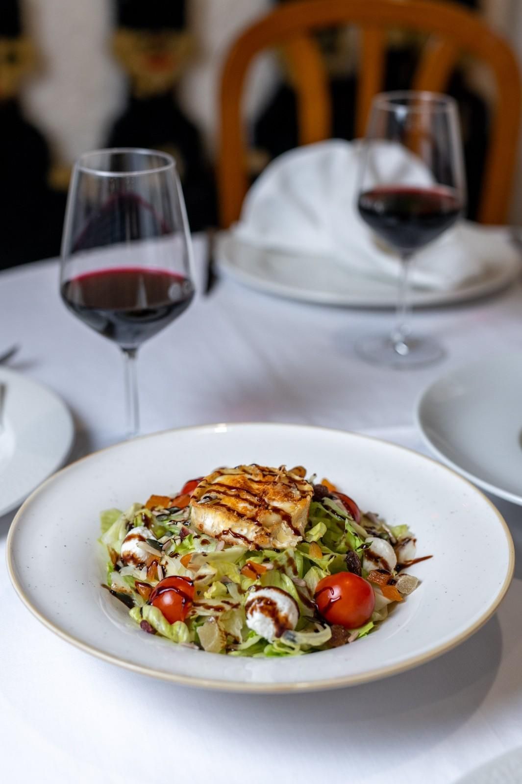 Plato blanco de ensalada sobre la mesa de un restaurante con dos copas de vino tinto y una servilleta doblada.