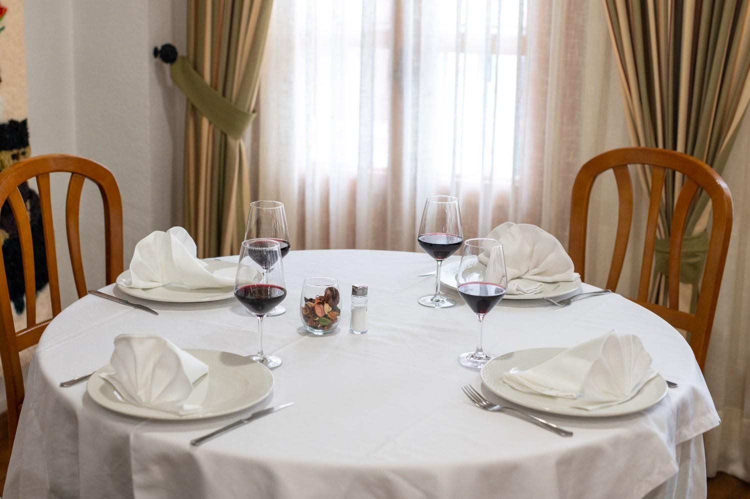 Coloca la mesa del comedor con platos blancos, copas de vino y servilletas dobladas junto a una ventana luminosa.