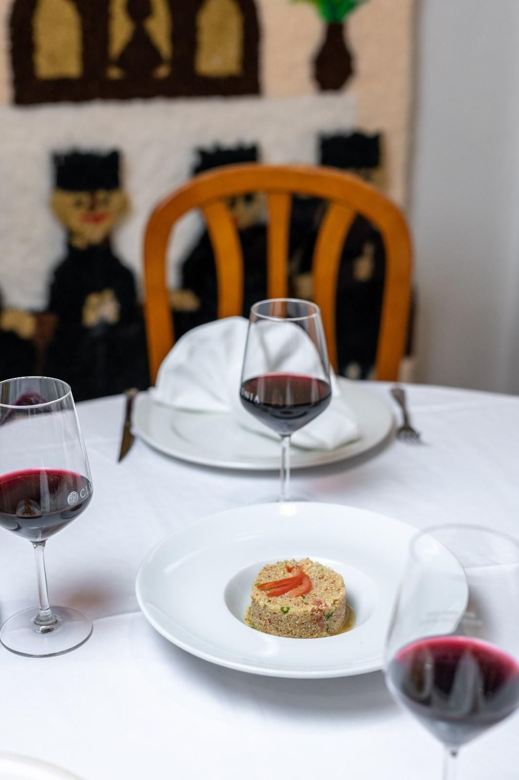 Mesa de restaurante puesta con platos blancos, copas de vino tinto y un aperitivo emplatado en primer plano.