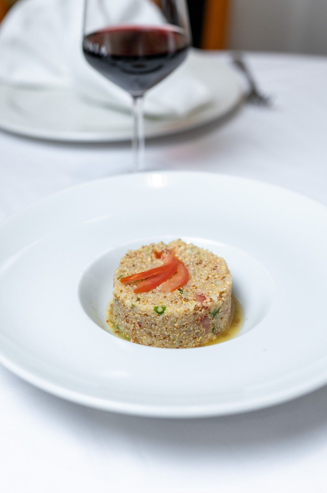 Un plato de cereales con verduras sobre un mantel blanco, con una copa de vino tinto al fondo.