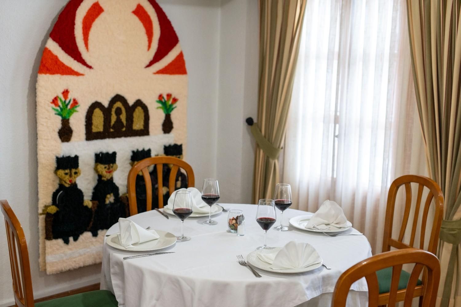 Mesa de comedor para cuatro personas junto a una chimenea blanca y cortinas que dejan pasar la luz del sol en una habitación acogedora.