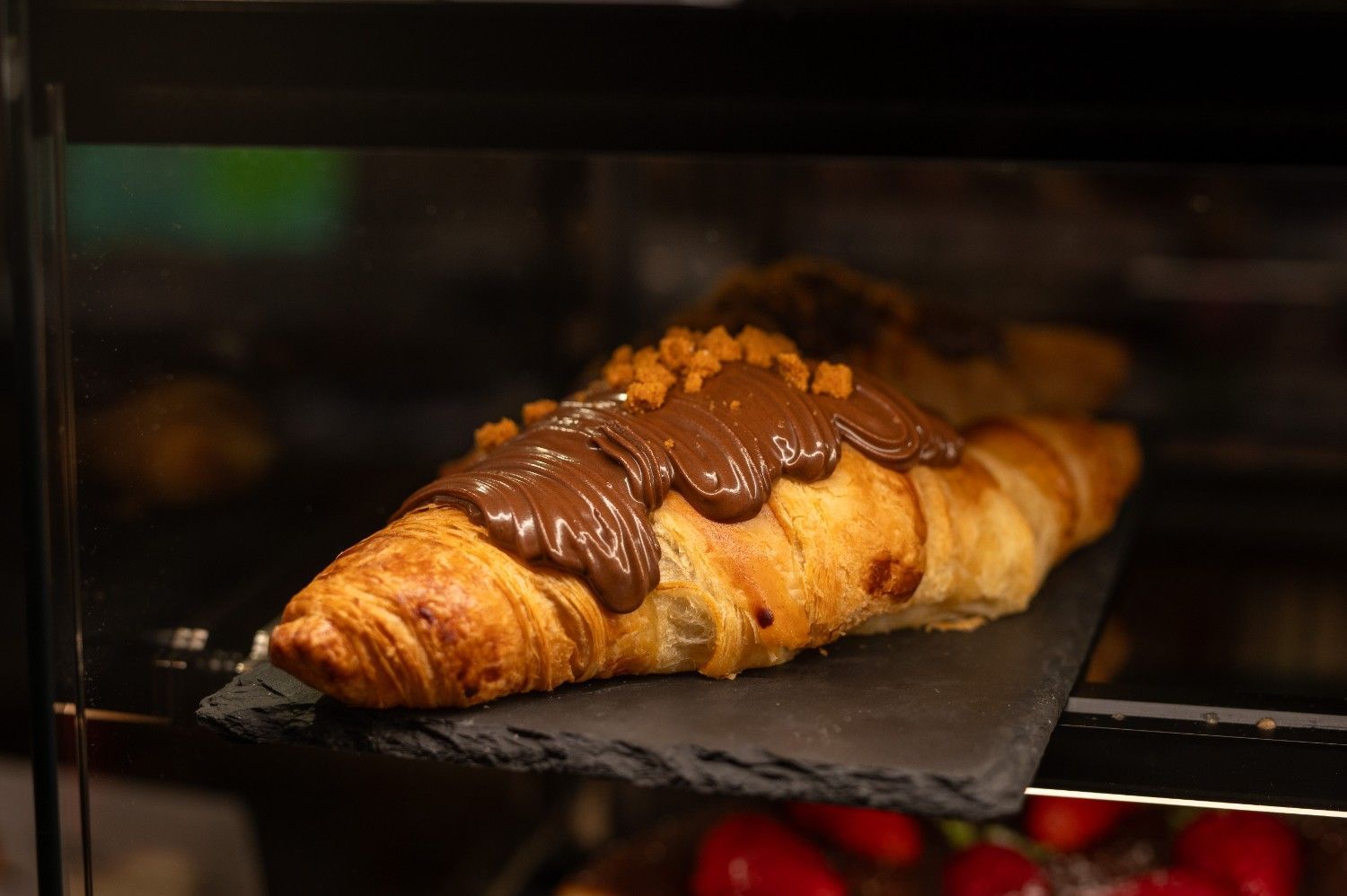 Croissant bañado en chocolate sobre una pizarra negra en una vitrina de panadería.