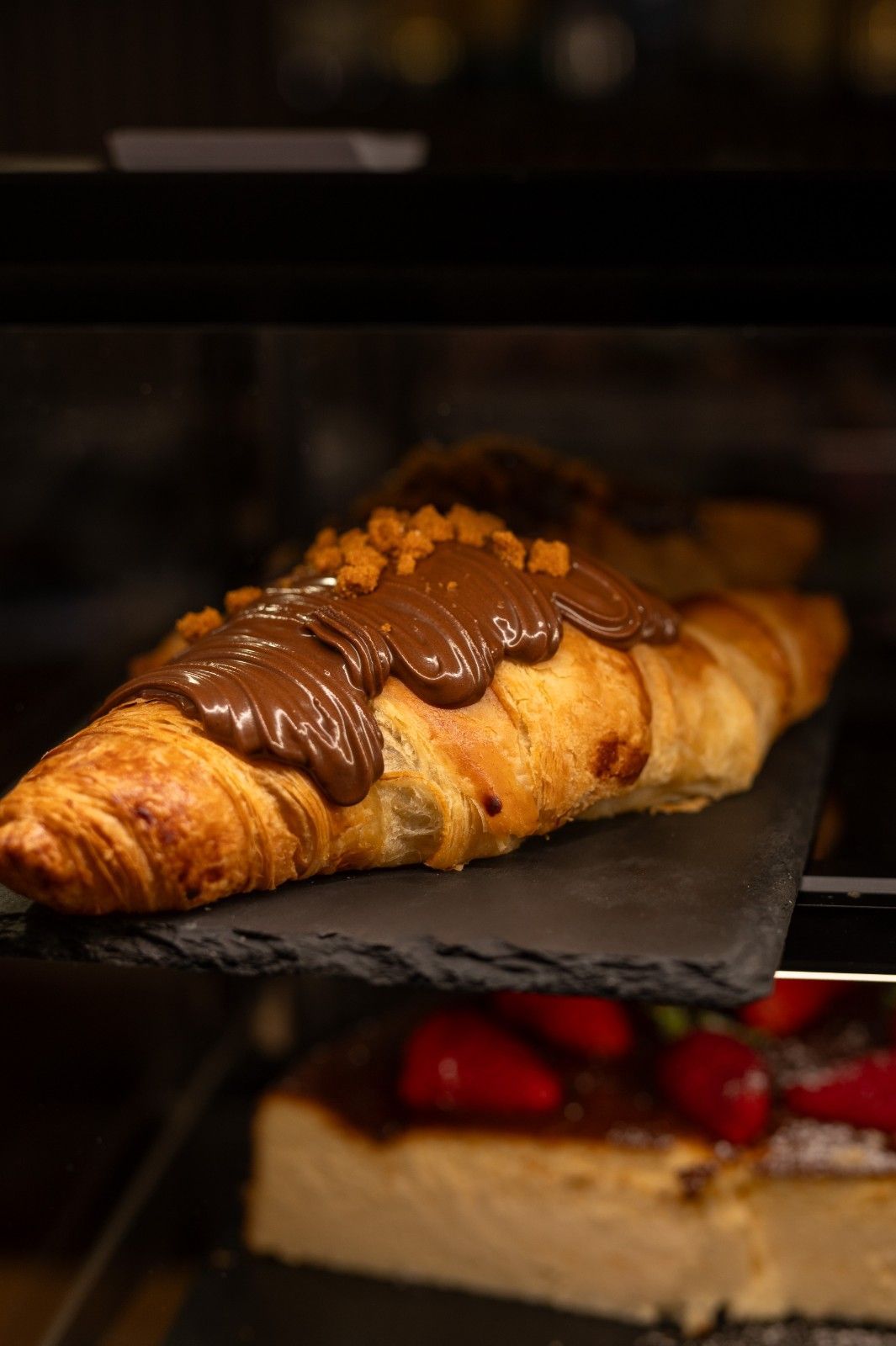 Un cruasán bañado en chocolate sobre una estantería de panadería, con una porción de postre y fresas debajo.