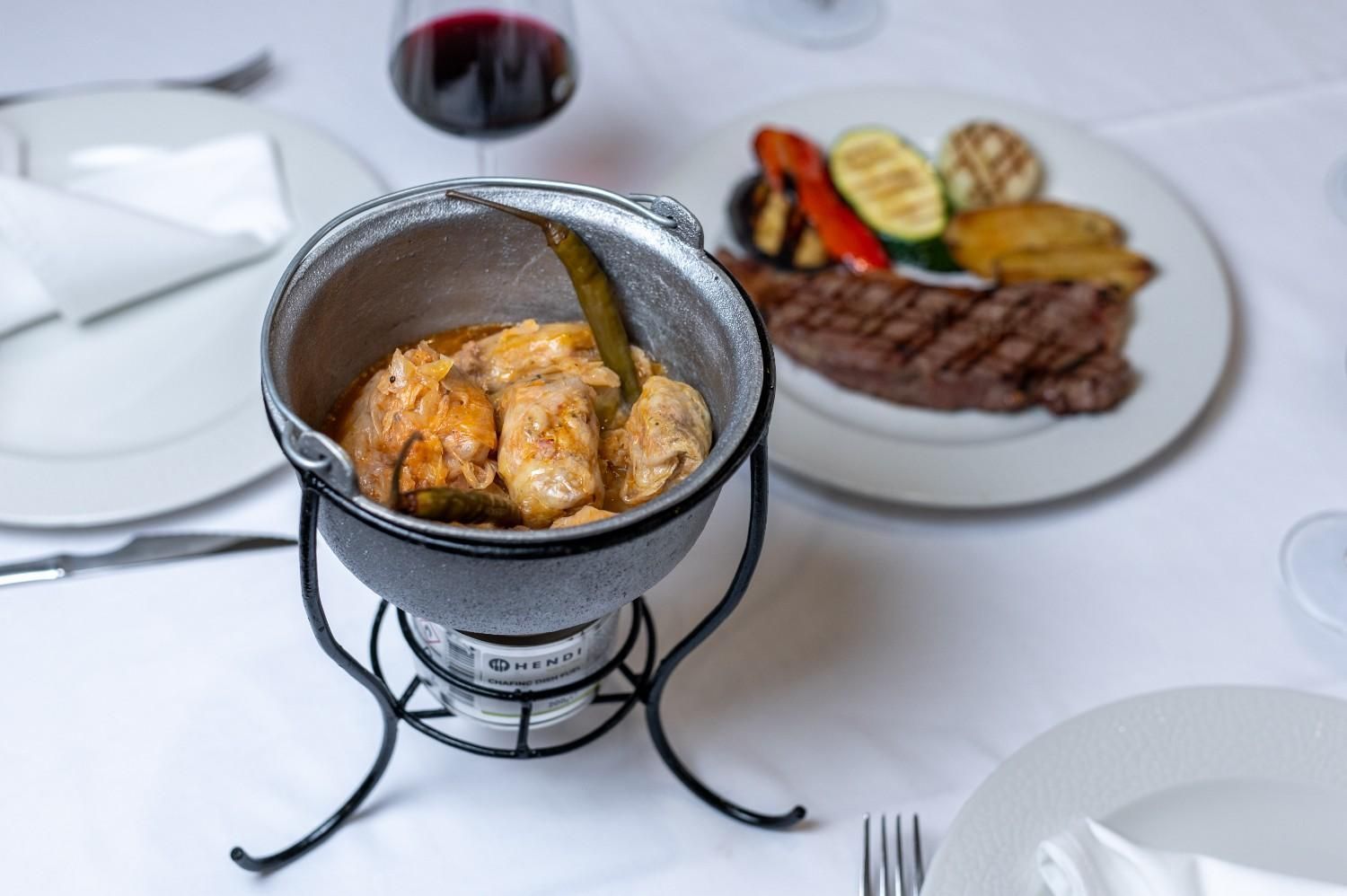 Elegante cena de bistec emplatado con verduras asadas y una olla de hierro fundido con comida sobre un mantel blanco.