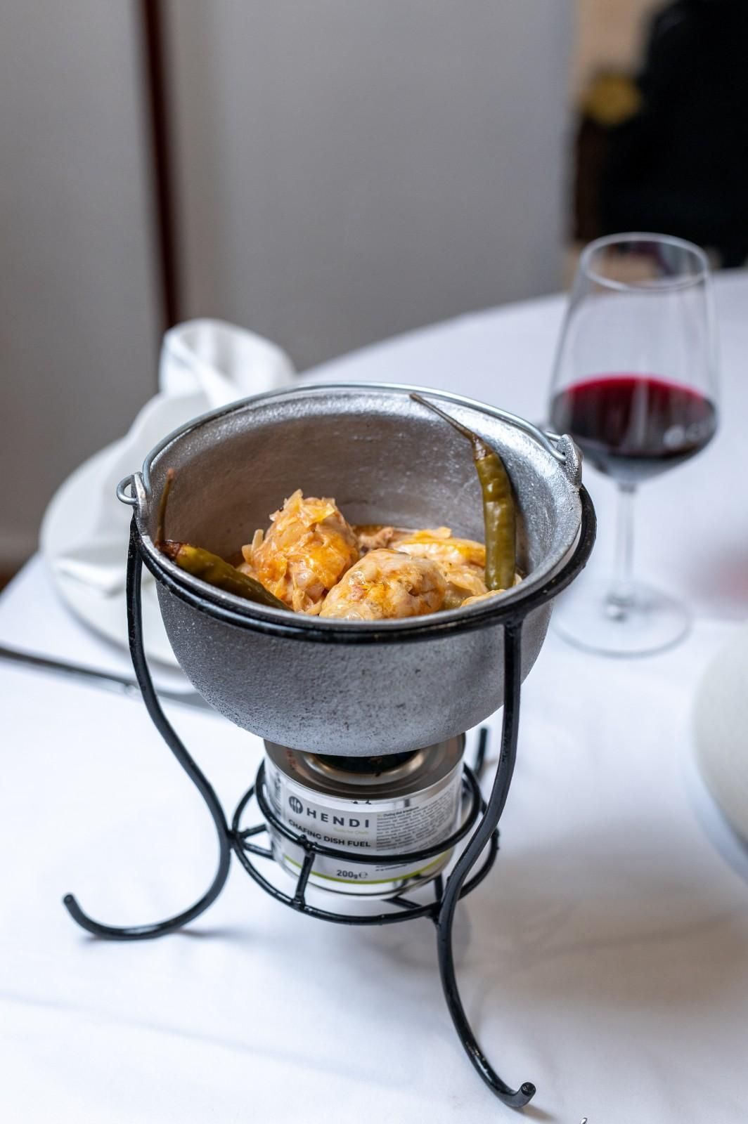 Una pequeña olla de fondue de mesa con queso derretido sobre una mesa de restaurante cubierta con un mantel blanco, junto a una copa de vino tinto.