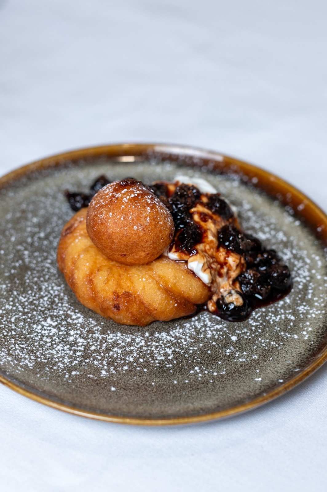 Pastel de postre espolvoreado con azúcar glas sobre un plato gris, cubierto con una fruta redonda y salsa oscura.