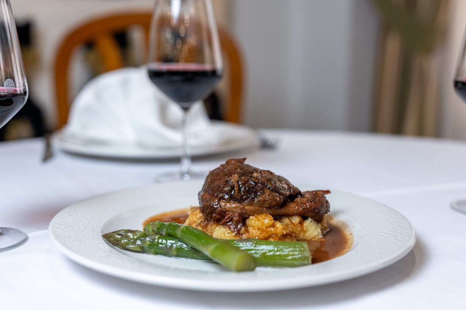 Plato gourmet con espárragos y carne sobre una mesa blanca, con copas de vino al fondo.