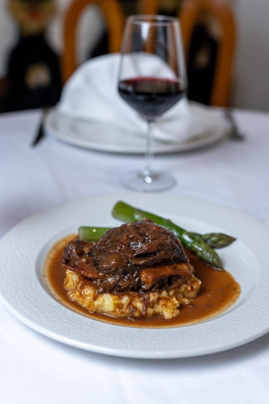 Carne estofada emplatada con verduras y salsa sobre una mesa blanca, con una copa de vino tinto detrás.