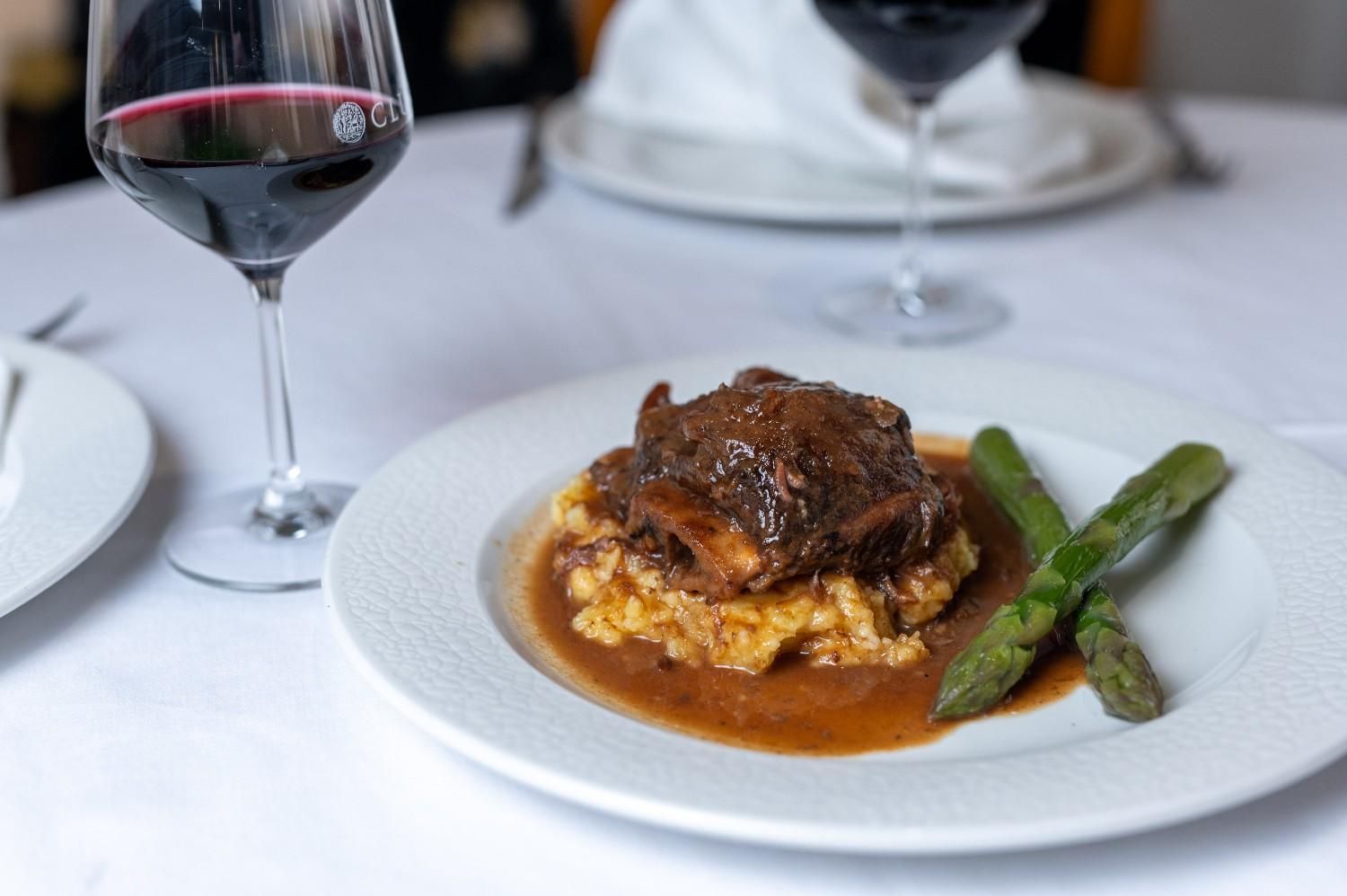 Carne emplatada con salsa y espárragos sobre una mesa blanca junto a copas de vino tinto.