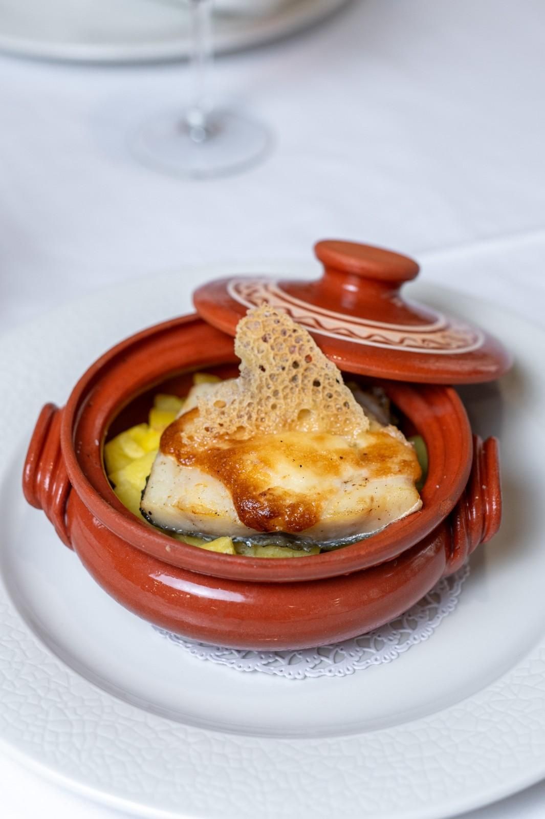 Cazuela pequeña de terracota sobre un plato blanco, cubierta con queso tostado y una guarnición crujiente.