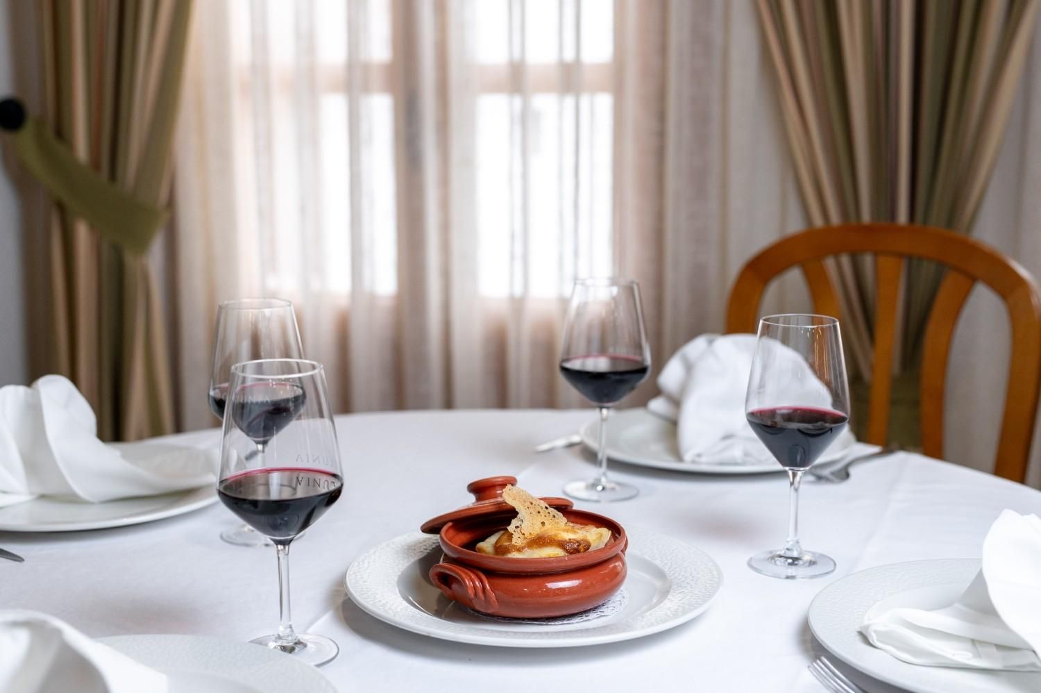 Mesa elegantemente puesta con copas de vino y un postre emplatado en un luminoso restaurante junto a una ventana.