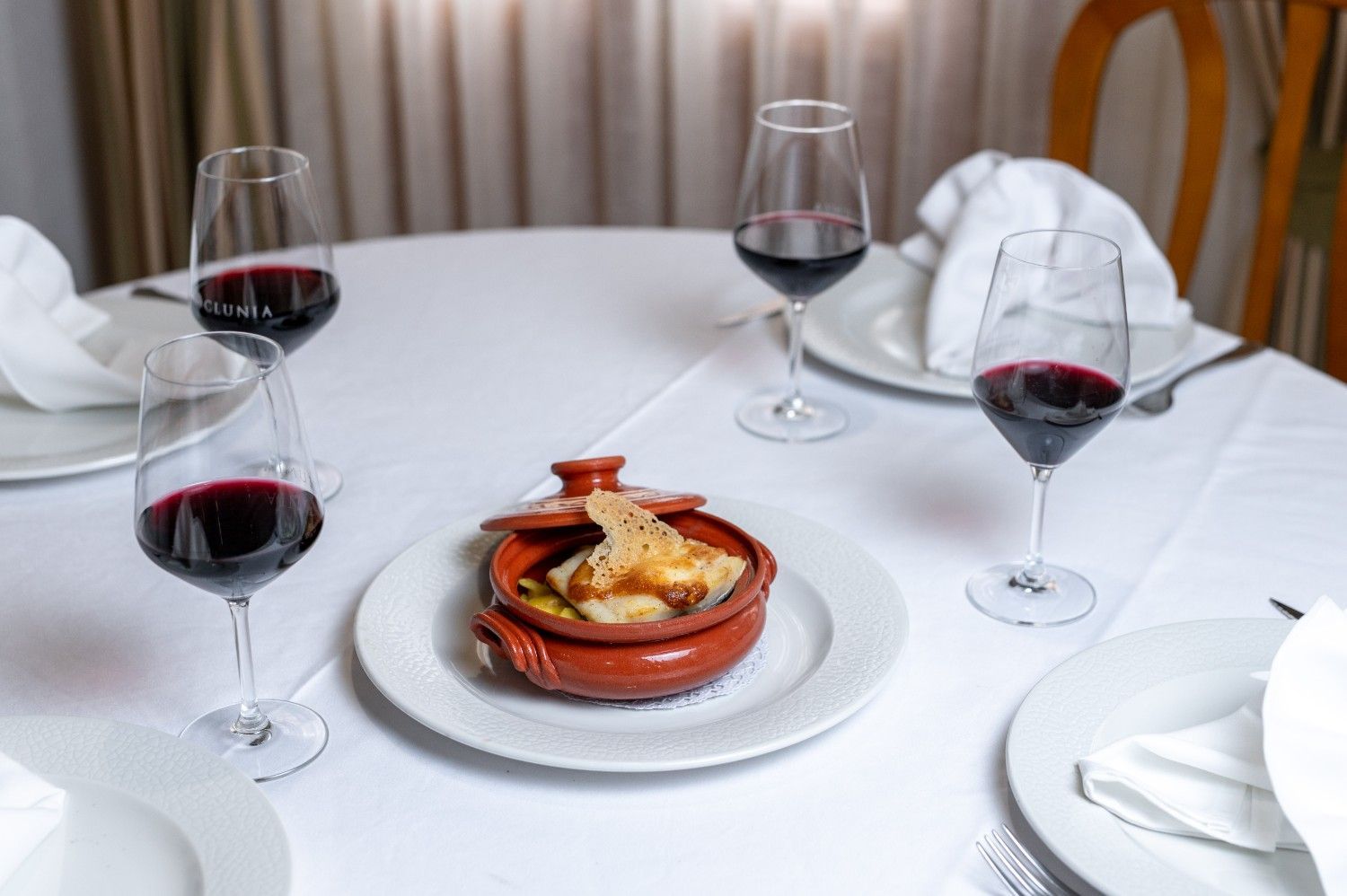 Elegante mesa de comedor con copas de vino tinto y una fuente de terracota con comida sobre un mantel blanco.