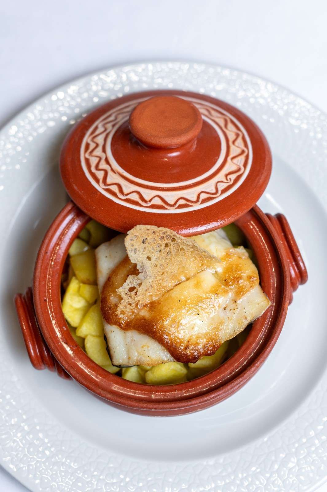 Cacerola de barro con tapa que contiene verduras y pan sobre un plato blanco.