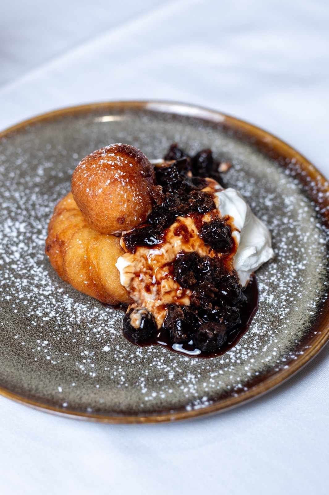 Plato de postre con buñuelos, crema batida y salsa de frutos rojos espolvoreada con azúcar glas.
