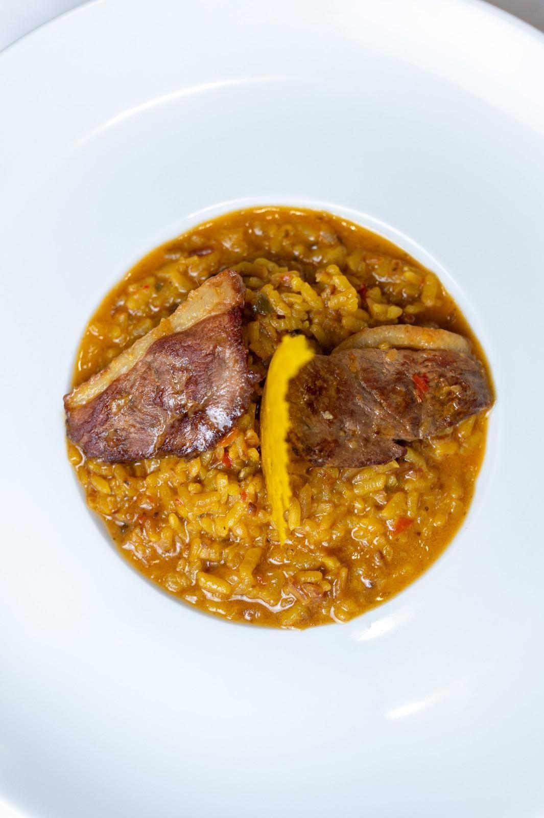 Un pequeño tazón de estofado de arroz naranja cubierto con dos trozos de carne dorada sobre un plato blanco.