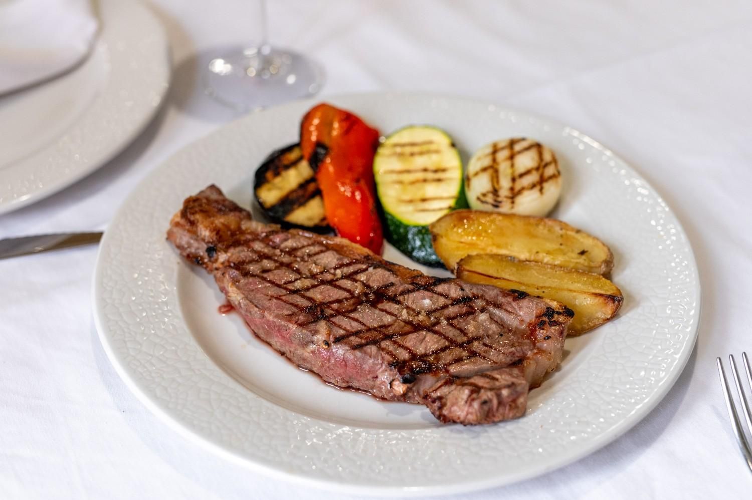 Filete a la parrilla con verduras asadas en un plato blanco en una mesa de comedor formal.