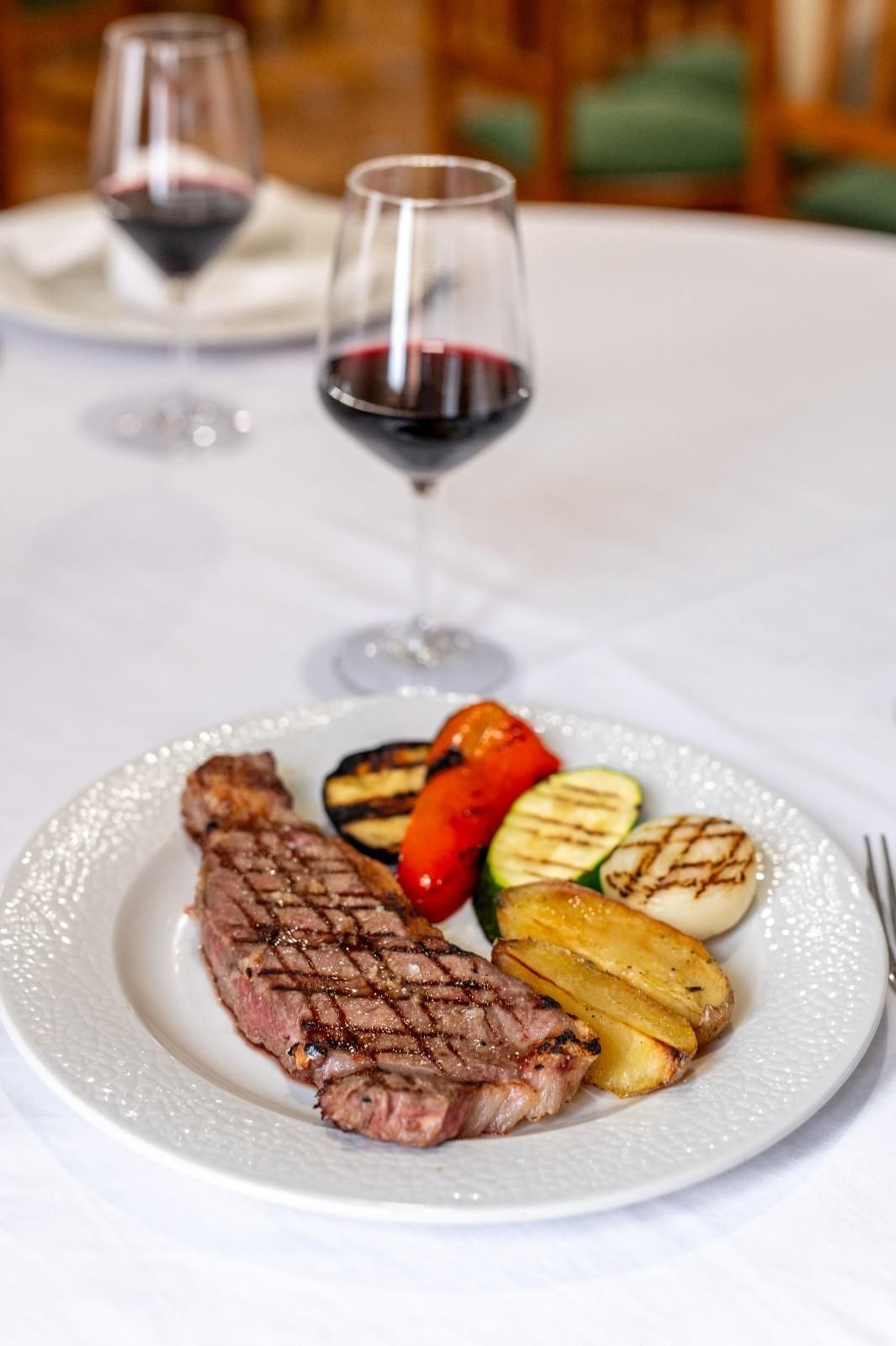 Filete a la parrilla y verduras en un plato blanco con dos copas de vino tinto en una mesa.