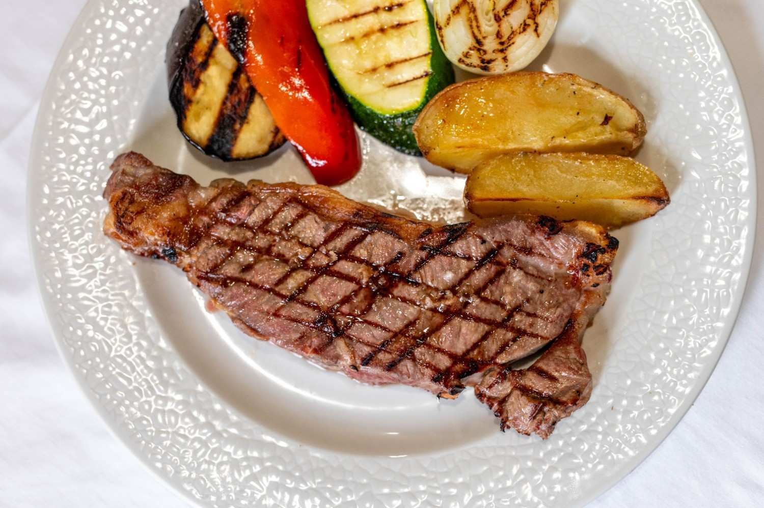 Filete a la parrilla con verduras asadas y patatas gajo en un plato blanco.