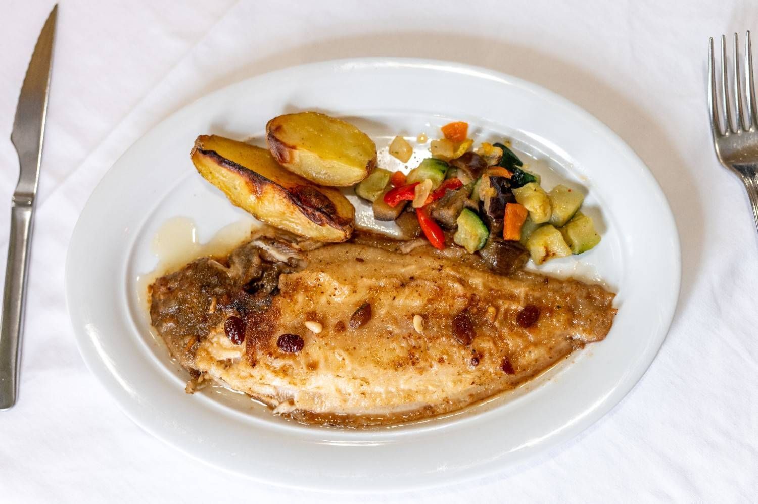 Pescado al horno emplatado con patatas asadas y verduras variadas sobre un mantel blanco.