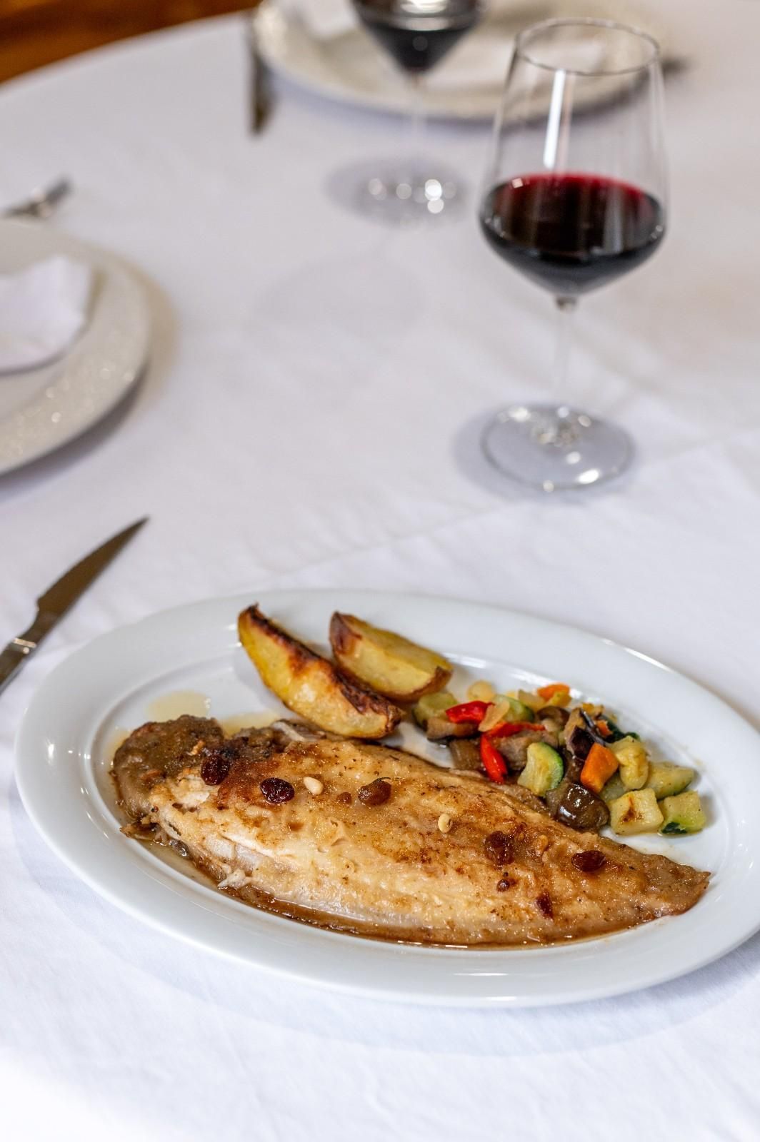 Pescado a la parrilla con verduras asadas en un plato blanco junto a una copa de vino tinto en una mesa.