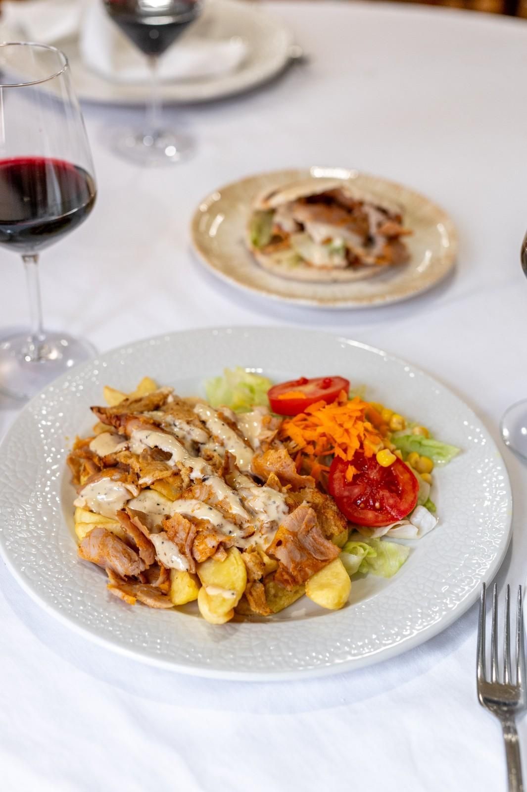 Plato gourmet servido en una mesa blanca con una copa de vino tinto y un pequeño aperitivo al fondo.