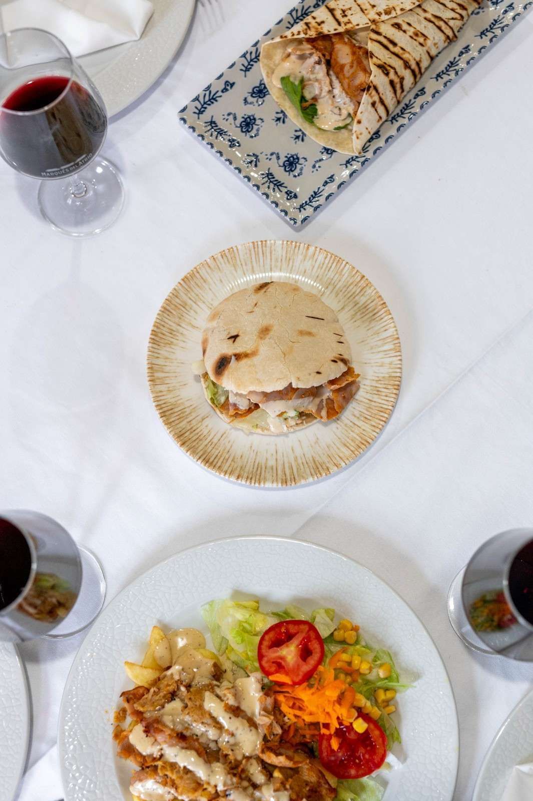 Vista aérea de una mesa con sándwiches de pita, ensalada y copas de vino tinto.