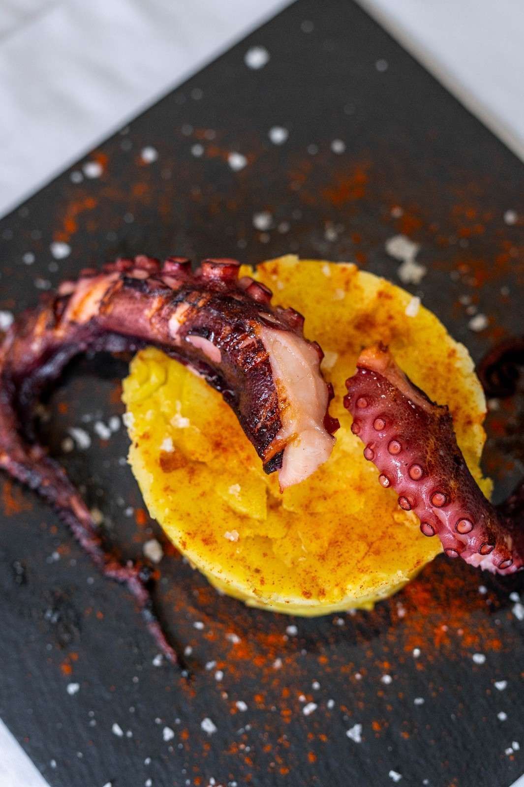Pulpo a la parrilla sobre salsa amarilla, espolvoreado con condimento rojo en un plato oscuro.
