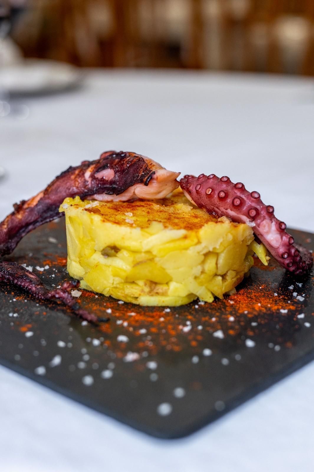 Tentáculo de pulpo cubriendo un plato amarillo en capas sobre un plato de pizarra negra.