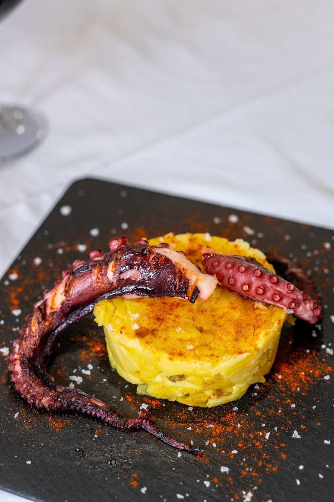 Tentáculo de pulpo a la parrilla sobre una torta de patata dorada en un plato negro, espolvoreado con condimento rojo.