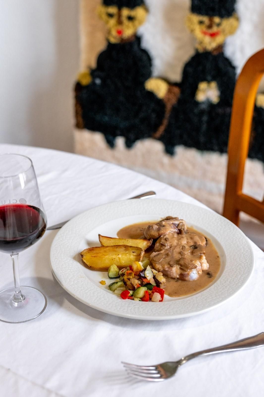 Una copa de vino y un plato servido sobre un mantel blanco, con un cuadro borroso al fondo.