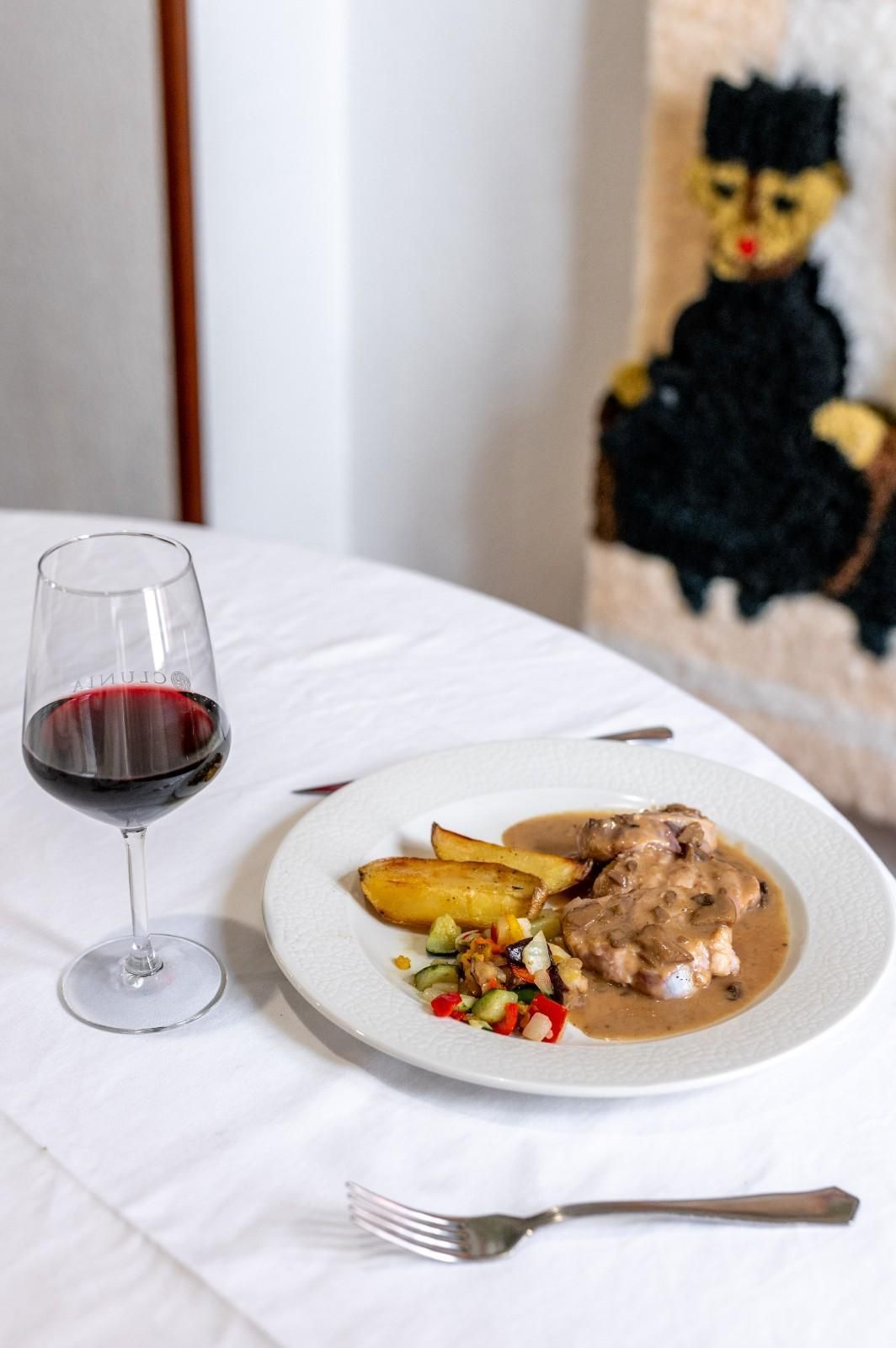 Copa de vino y plato servido sobre un mantel blanco, con un tenedor y un cuadro decorativo al fondo.