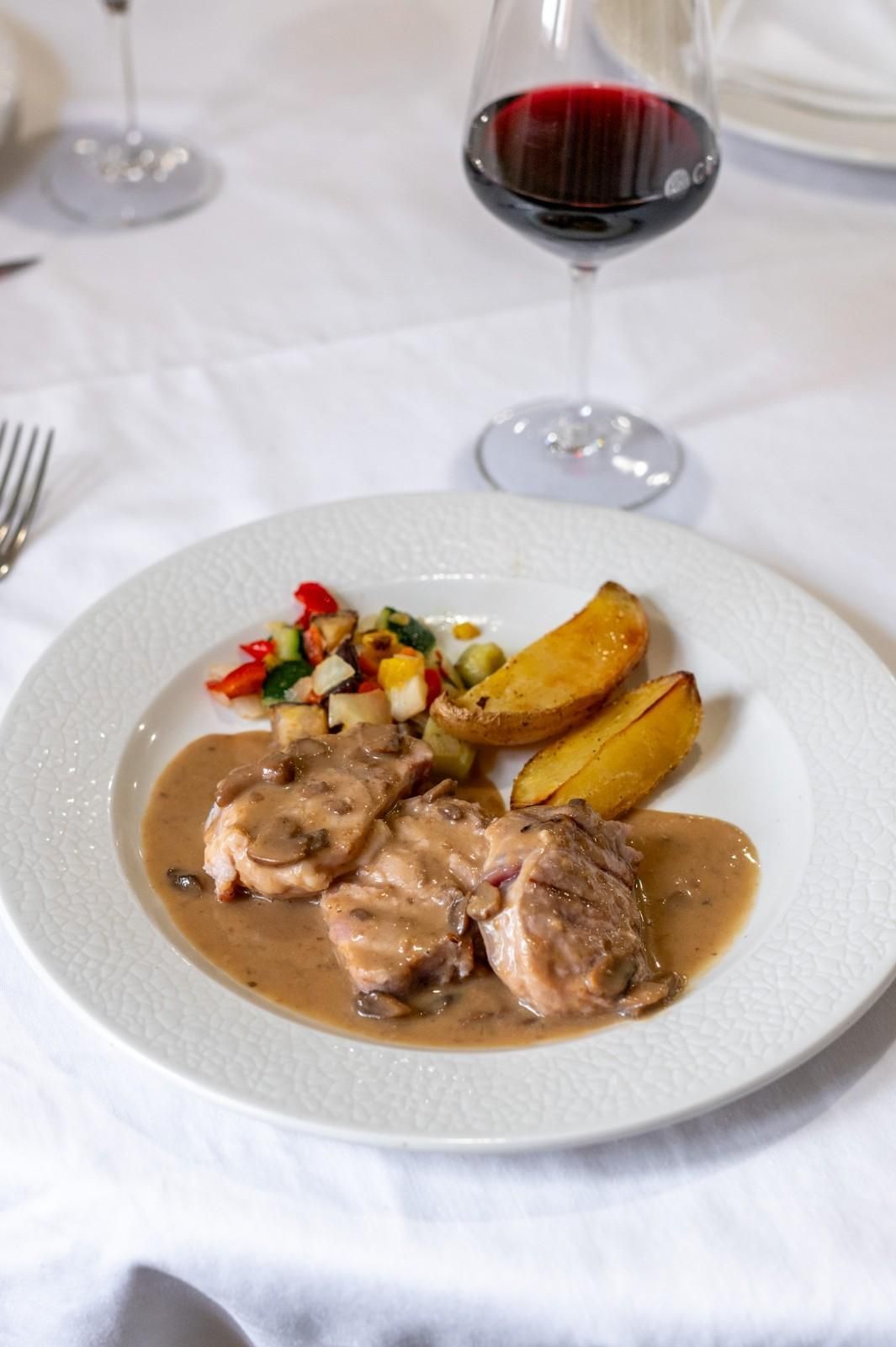 Plato de carne en salsa marrón con patatas asadas y guarnición colorida, acompañado de una copa de vino tinto.