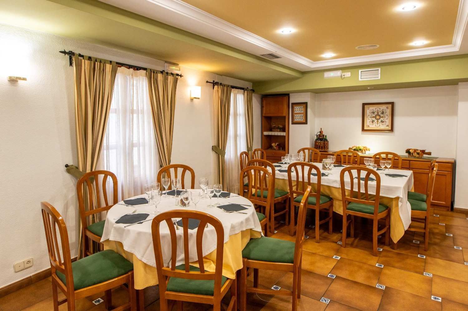 Comedor con sillas de madera, mesas redondas e iluminación cálida amarilla, visible a través de grandes ventanales con cortinas.