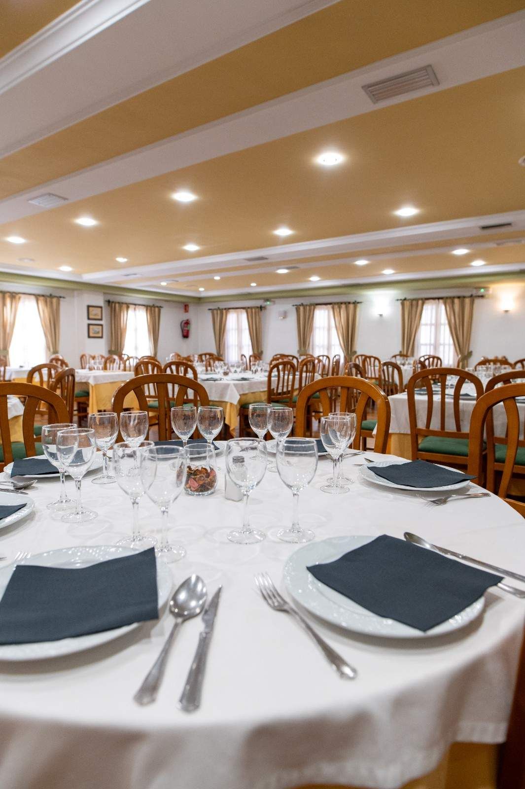 Mesa de comedor preparada en un luminoso salón de banquetes de restaurante con sillas de madera y manteles blancos.