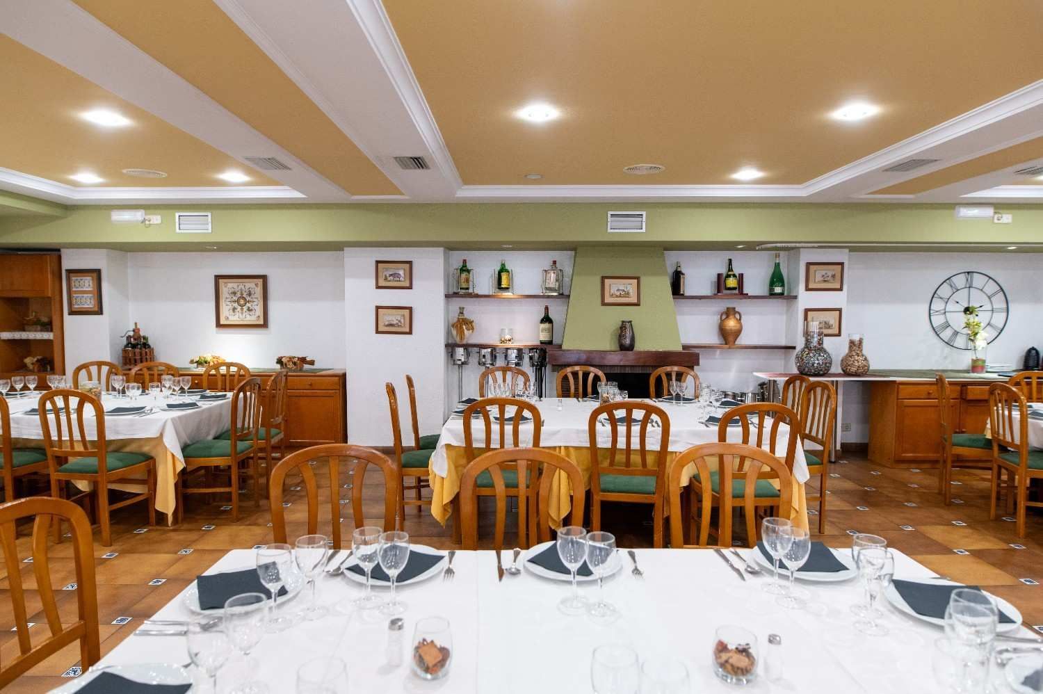 Comedor luminoso con mesas puestas, sillas de madera y estantes de pared bajo luces de techo cálidas.