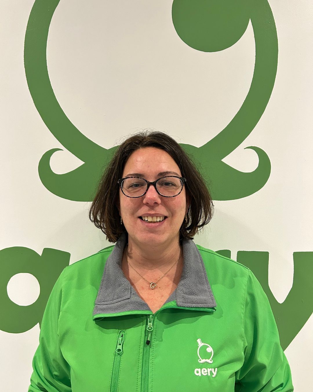 Une femme portant des lunettes et une veste verte sourit devant un logo vert.