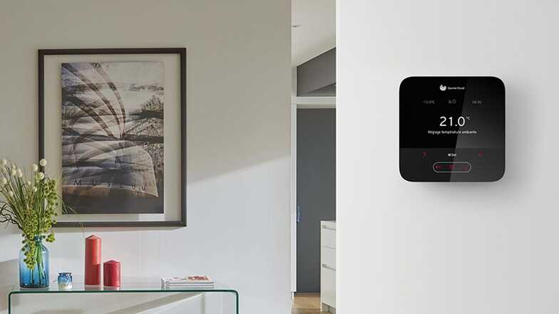 Thermostat intelligent fixé au mur blanc, affichant la température ; œuvre d'art et table décorative dans la pièce.