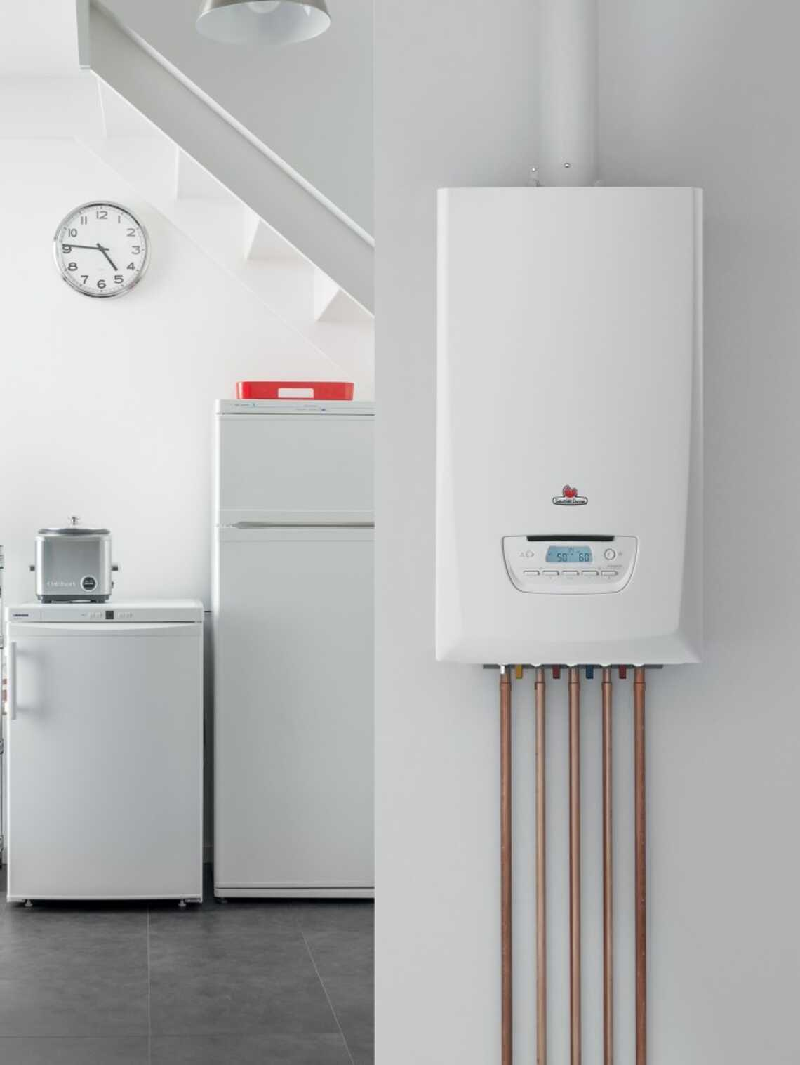 Chaudière murale blanche avec tuyaux en cuivre, horloge, réfrigérateur et petits appareils électroménagers dans une pièce.