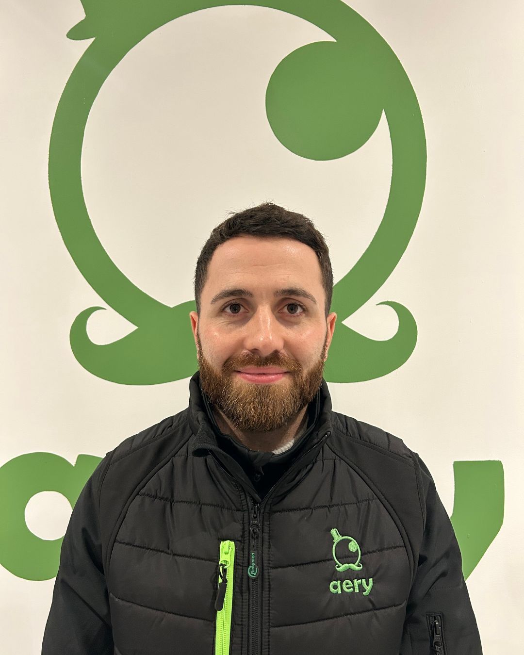 Un homme barbu portant une veste noire à fermeture éclair verte se tient devant un logo vert sur un mur blanc.