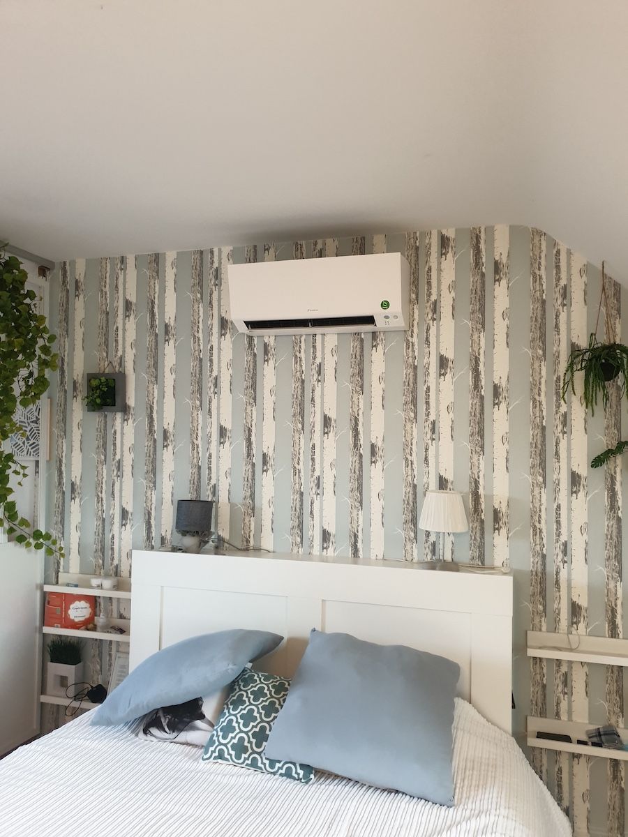 Chambre avec tête de lit blanche, climatisation, papier peint à motifs et plantes suspendues.