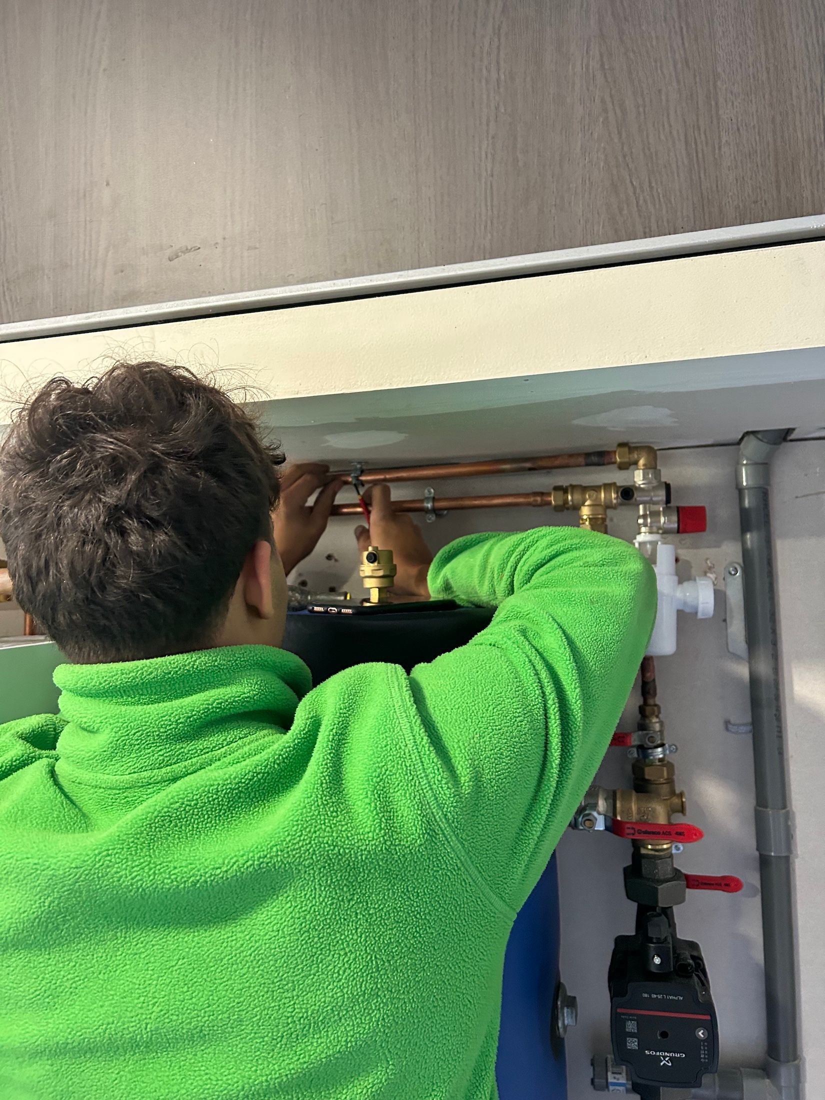 Une personne en veste verte travaille sur des tuyaux et des vannes en cuivre, fixés au mur.