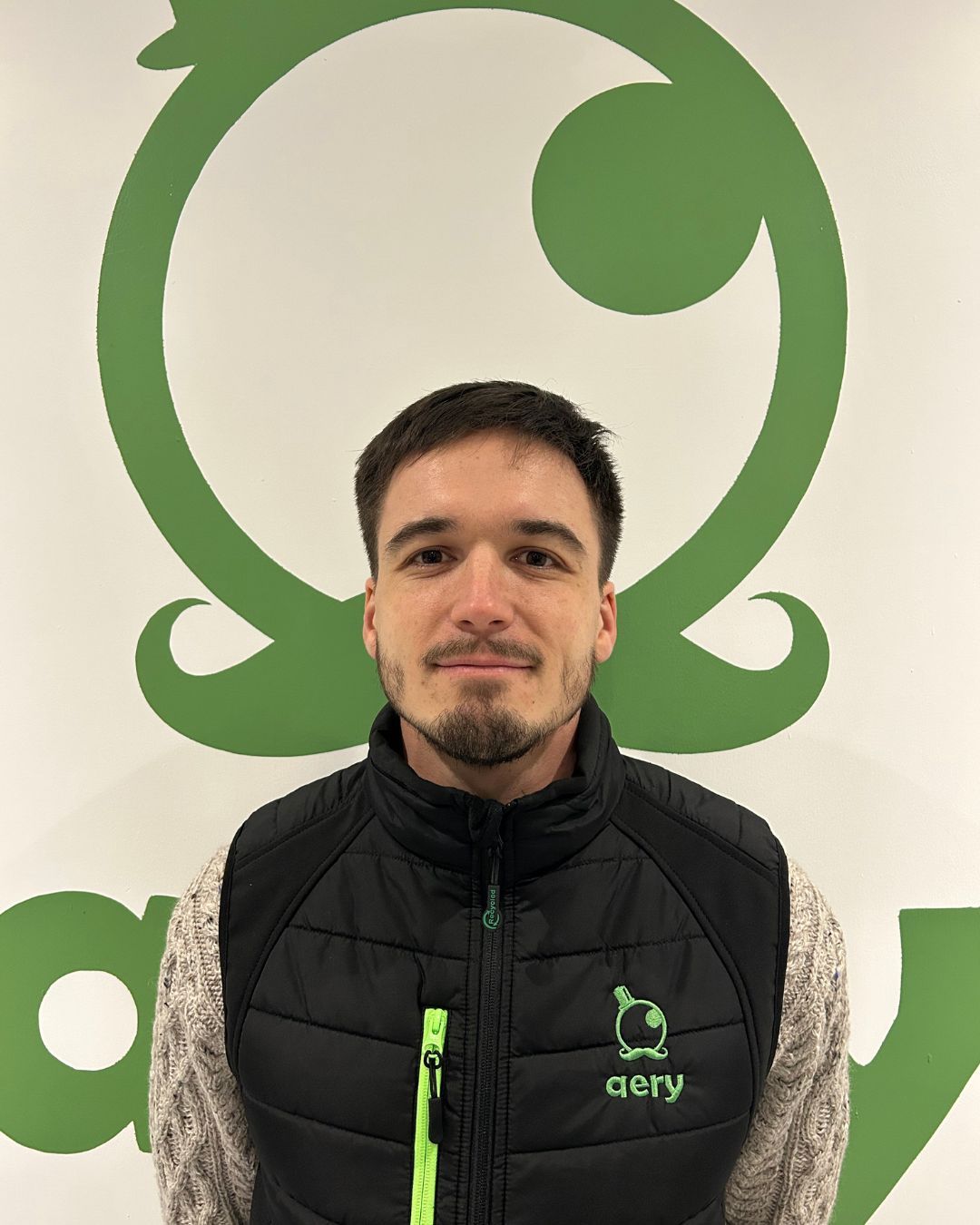 Un homme portant un gilet noir et gris se tient devant un logo vert sur un mur blanc.